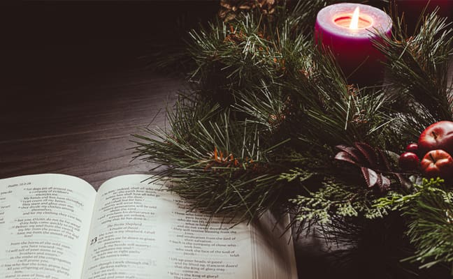 An open Bible resting on a dark wooden surface, surrounded by a pine wreath and lit purple candles, creating a warm and reflective Advent atmosphere.